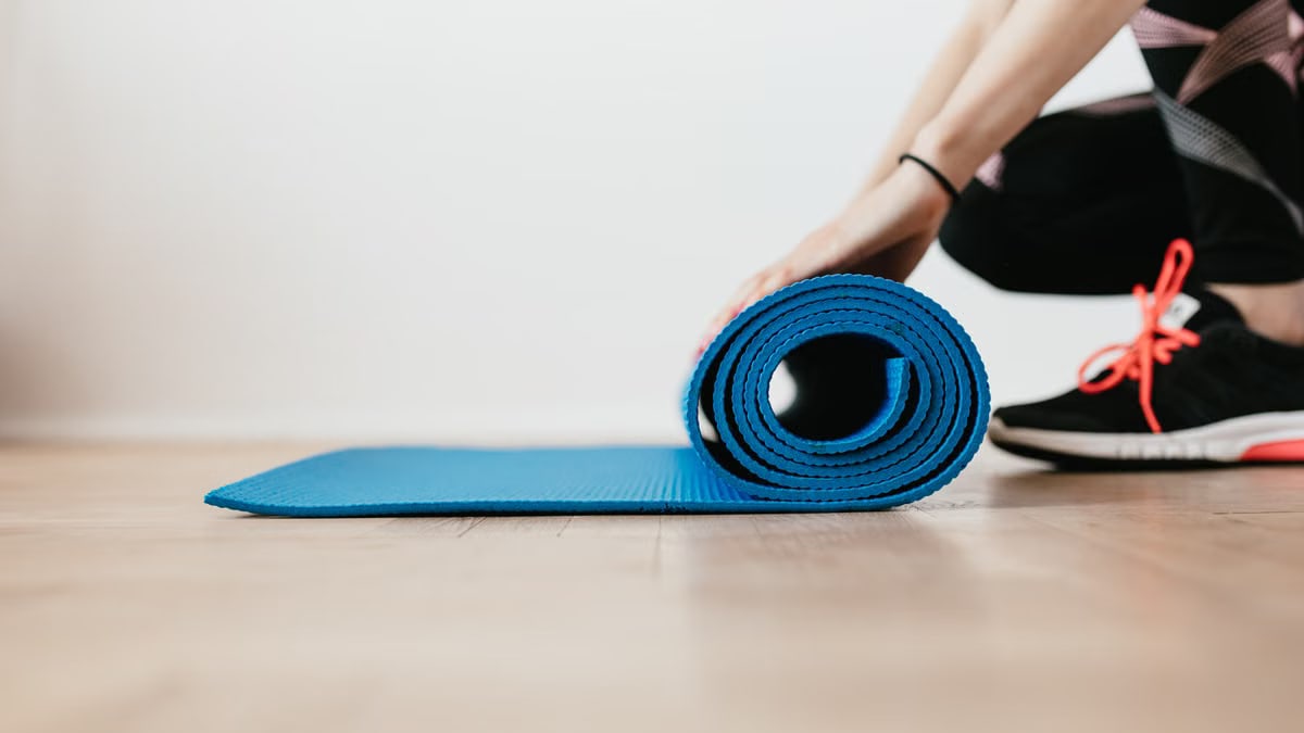 Side view of unrecognizable female athlete in black leggings and sneakers unfolding sport mat for fitness training indoors