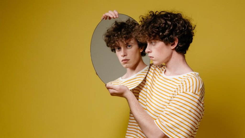 Studio portrait of a young man holding a mirror against a yellow background.